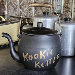 May include: Black ceramic cookie kettle with a gold-colored handle and the words "Kookie Kettle" in gold lettering. The kettle has a spout and a lid. Other metal canisters are in the background.
