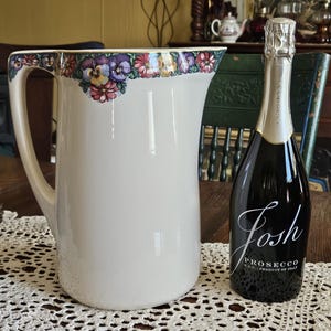May include: A white ceramic pitcher featuring a colorful floral border of pansies and daisies along the top rim and handle, positioned next to a dark glass bottle of Josh Prosecco with white text on the label indicating the brand and product type.