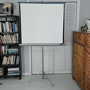 May include: A portable projector screen with a white rectangular surface and a silver metal housing, mounted on a collapsible silver tripod stand. The screen is fully extended vertically, featuring a thin blue top border and a silver bottom bar.