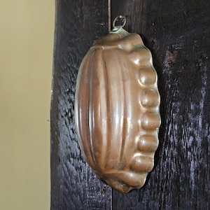 May include: A vintage copper mold, oval-shaped with a ribbed design on one side and a series of rounded bumps on the other. It has a loop at the top for hanging. The mold is a warm, antique copper color.