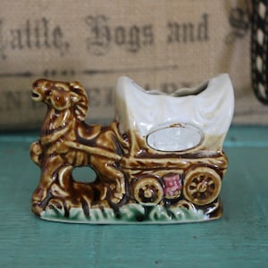 May include: A brown and white ceramic figurine of a horse-drawn covered wagon. The wagon has a white canvas top and brown wheels. The horse is brown and has a white mane and tail.