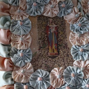 May include: A framed image of a religious figure with a baby in their arms, surrounded by a border of handmade fabric flowers in shades of pink and blue.