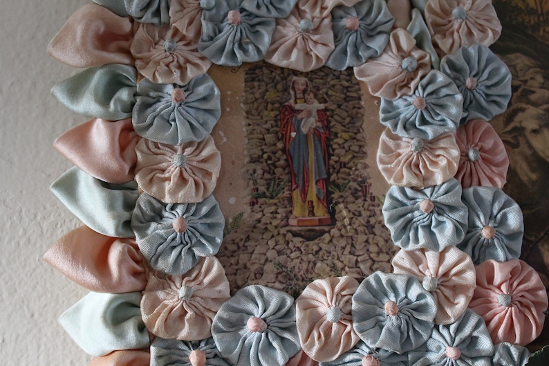 May include: A framed image of a religious figure with a baby in their arms, surrounded by a border of handmade fabric flowers in shades of pink and blue.