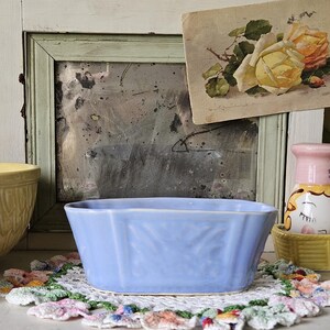 May include: A light blue ceramic planter with a rectangular shape and leaf pattern. The planter sits on a colorful, crocheted doily. A vintage postcard with yellow roses is in the background.