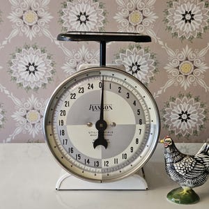 May include: A vintage Hanson utility scale with a round face, black needle, and white base, featuring a dial marked from 0 to 24 pounds. Beside the scale stands a small, detailed ceramic chicken figurine with black, white, and yellow painted accents.
