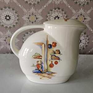 May include: Cream-colored ceramic pitcher with a lid and a curved handle. The pitcher features a painted scene of a person in a sombrero, with shelves of pottery and food. The background is a floral wallpaper.