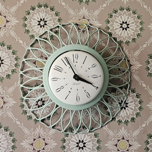 May include: A round wall clock with a mint green metal sunburst frame and a white face. The clock features black stylized numerals, black hour and minute hands, and the word Sessions printed in black script in the center of the white dial.