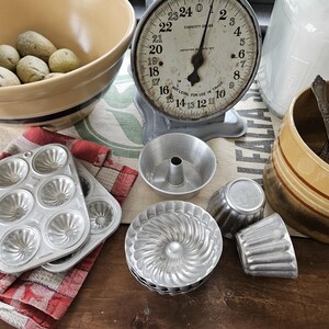 May include: A collection of vintage baking tools and ingredients. Includes a large yellow and blue striped bowl, a scale, metal bundt cake molds, and a red and white checkered cloth. Potatoes are in the bowl.