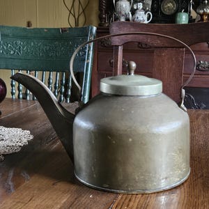 May include: An antique brass kettle with a rounded body, a spout, and a handle. The lid is flat with a small knob. The kettle sits on a wooden table. The overall aesthetic suggests a vintage or rustic theme.