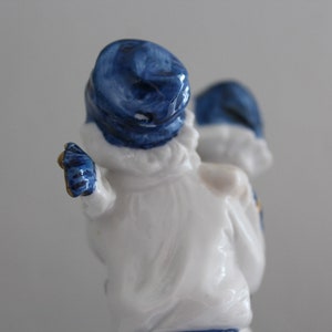 May include: A white porcelain figurine of two people wearing blue hats. The figurine is standing and the person at the front is holding out their arm.