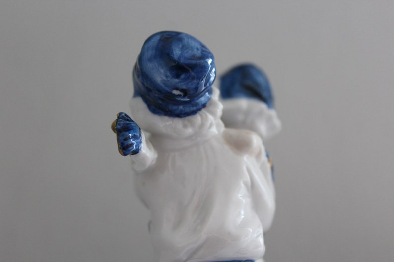 May include: A white porcelain figurine of two people wearing blue hats. The figurine is standing and the person at the front is holding out their arm.