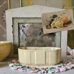 May include: A still life arrangement featuring a pale yellow rectangular ceramic dish, a round yellow bowl, and a vintage postcard with rose illustrations. A white ceramic chicken figurine and a colorful doily complete the scene.