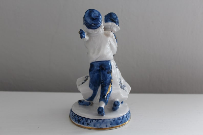 May include: A porcelain figurine of a couple dancing, painted in blue and white with gold accents. The man is wearing a blue hat and the woman is wearing a white dress with blue trim.