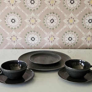 May include: A set of dark gray ceramic tableware, including two teacups with saucers, a large plate, and a smaller plate. The background features a patterned wallpaper with floral designs in shades of beige, white, and green.