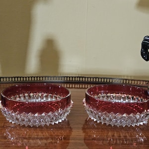 May include: Two ruby red and clear crystal bowls with a starburst design. The bowls have a cut crystal base with a decorative rim. A silver cocktail shaker and a black horse statue are in the background.