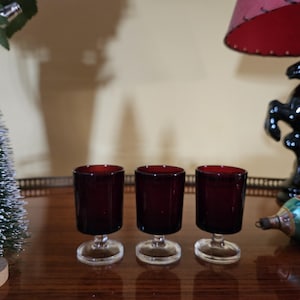 May include: Three small, clear glass shot glasses with a red, opaque base. The glasses are arranged in a row on a wooden surface.