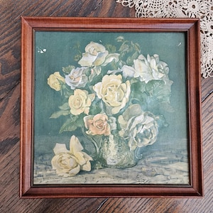 May include: A framed floral print featuring a bouquet of white, cream, and peach roses in a decorative silver pitcher. The artwork is set against a teal background and framed in a dark brown wooden frame. The overall theme is vintage floral art.