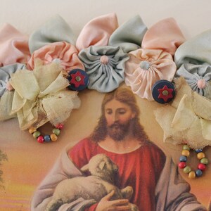 May include: A vintage religious image of Jesus holding a lamb, framed with a colorful fabric flower garland. The garland is made of pastel pink, blue, and yellow fabric flowers.