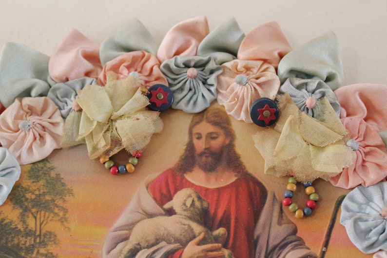 May include: A vintage religious image of Jesus holding a lamb, framed with a colorful fabric flower garland. The garland is made of pastel pink, blue, and yellow fabric flowers.