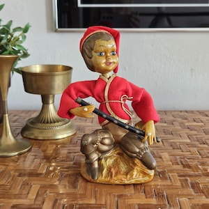 May include: A vintage figurine of a musician in a red outfit with gold trim, holding a black flute. The figure has a wooden head and is seated on a gold-colored base. Decorative brass vases are in the background.