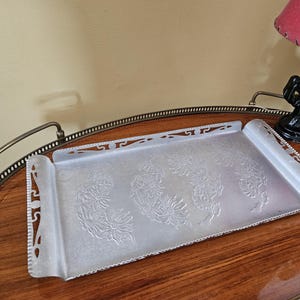 May include: A rectangular, silver-colored metal tray with embossed floral designs. The tray has decorative cut-out edges and is resting on a wooden table. A lamp with a red shade is visible in the background.