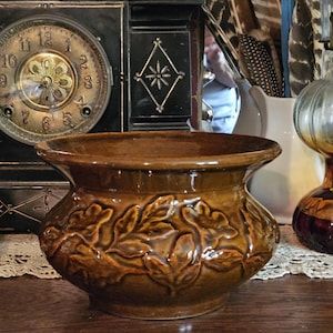 May include: A brown ceramic planter with a detailed leaf pattern. The planter sits on a wooden surface, with a vintage clock and other decorative items in the background. The planter is approximately 8 inches in diameter.