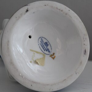 May include: The underside of a white porcelain figurine with a blue and gold design. The figurine has a small hole in the base and a gold sticker with the text "Goebel Germany" and a rose.
