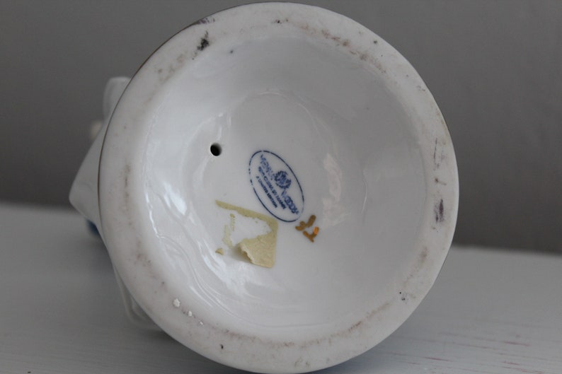 May include: The underside of a white porcelain figurine with a blue and gold design. The figurine has a small hole in the base and a gold sticker with the text "Goebel Germany" and a rose.