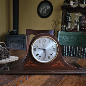May include: A dark brown wooden mantel clock with a curved top and a wide base. The clock face is circular with a silver background, black Arabic numerals, and a gold-toned bezel. Two decorative leaf-shaped inlays flank the clock face.