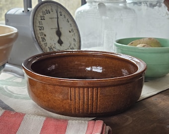 May include: A brown ceramic bowl with a ribbed design, set on a wooden surface. A vintage scale and other bowls are in the background. The bowl is approximately 10 inches in diameter.