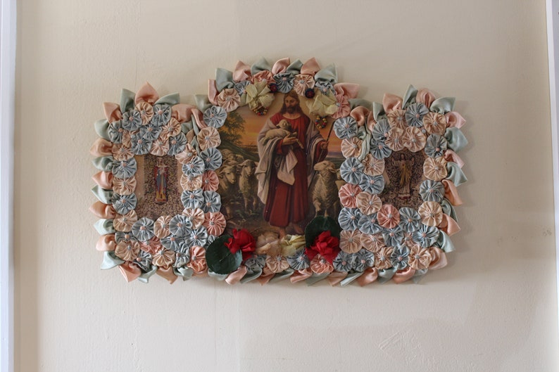 May include: A framed religious image of Jesus carrying a lamb, surrounded by a decorative border of fabric flowers in shades of pink, blue, and green.