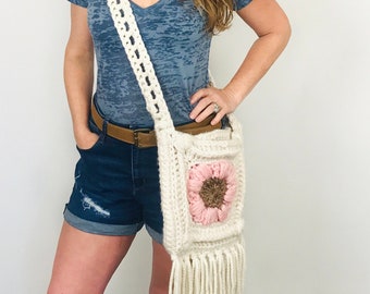 May include: A white crocheted crossbody bag with a pink and brown flower design and fringe. The bag is worn by a person wearing a blue shirt and denim shorts.
