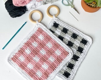 May include: Two crocheted pot holders hanging from wooden rings. One pot holder is pink and white with a polka dot pattern. The other pot holder is black and white with a checkered pattern.