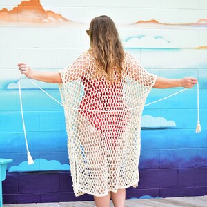 May include: A woman wearing a white crochet cover-up with tassels over a red swimsuit. The cover-up is a loose-fitting, open-front style with a long, flowing silhouette. The woman is standing in front of a blue and white wall with a mural of clouds.