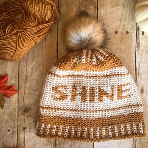 May include: A crocheted beanie hat in brown and white with the word "MINE" in brown letters. The hat has a fluffy brown pom pom on top.