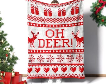 May include: A white and red knitted blanket with a festive design. The blanket features red gift boxes, geometric patterns, reindeer, snowflakes, and hearts. The words "OH DEER!" are prominently displayed in the center of the blanket.