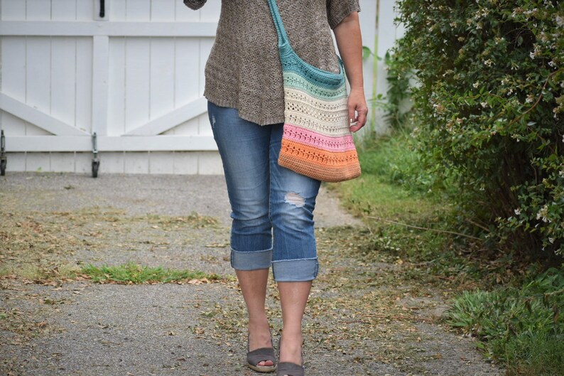 May include: A woman is wearing blue jeans and a brown sweater. She is carrying a multi-colored crochet tote bag with a long strap.