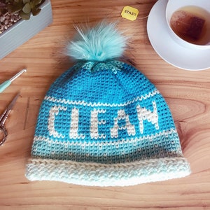 May include: A blue crocheted hat with a white pom pom and the word "CLEAN" in white yarn. The hat has a white and blue ombre effect.