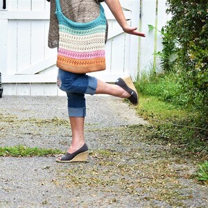 May include: A woman wearing blue jeans and a brown sweater is carrying a colorful crocheted tote bag. The bag has stripes of turquoise, white, pink, and orange.