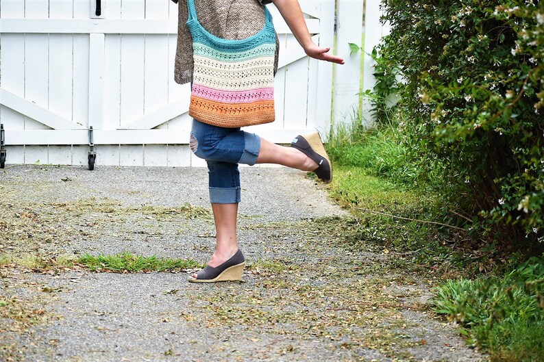 May include: A woman wearing blue jeans and a brown sweater is carrying a colorful crocheted tote bag. The bag has stripes of turquoise, white, pink, and orange.