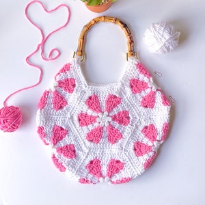 Op de afbeelding: Een witte gehaakte handtas met roze bloemenmotieven en een houten handvat.