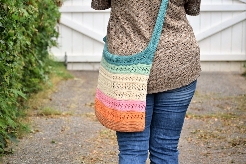 May include: A multi-colored crocheted tote bag with a long strap worn over the shoulder. The bag is made with a variety of colors, including teal, cream, pink, and orange.