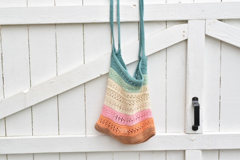May include: A crocheted tote bag with a rainbow color pattern. The bag is made with a light blue, light green, cream, pink, and orange yarn. The bag is hanging on a white wooden door.