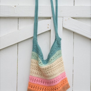 May include: A multi-colored crocheted tote bag with a teal, green, cream, pink, and orange color scheme. The bag is hanging on a white wooden fence.