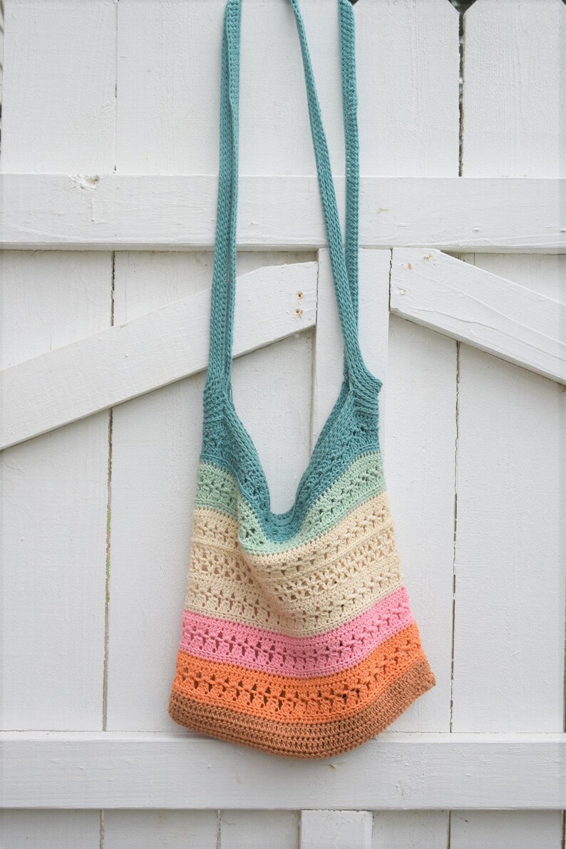May include: A multi-colored crocheted tote bag with a teal, green, cream, pink, and orange color scheme. The bag is hanging on a white wooden fence.