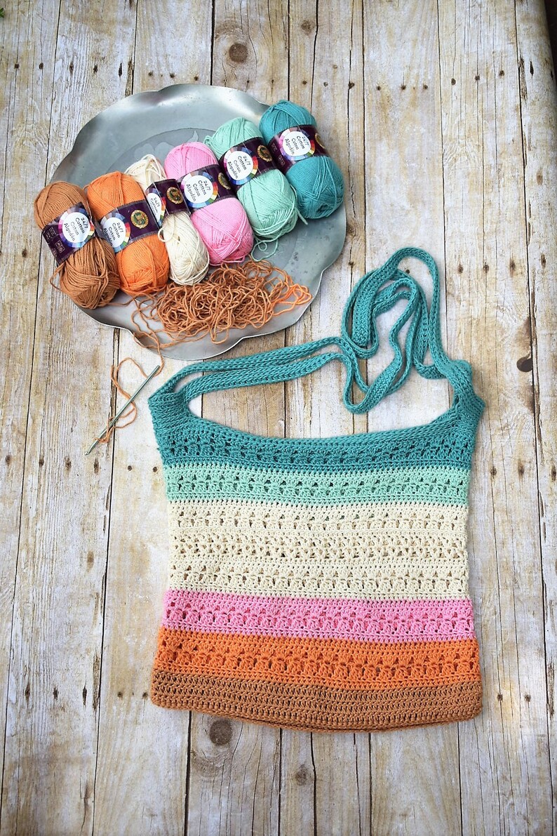 May include: A crocheted tote bag with a teal, green, cream, pink, orange, and brown striped design. The bag has two long teal straps. The bag is laying on a wooden surface with balls of yarn in a silver tray.