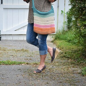 May include: A woman is wearing a pair of blue jeans and a brown sweater. She is carrying a multi-colored crochet tote bag with a teal, white, pink, and orange striped design.