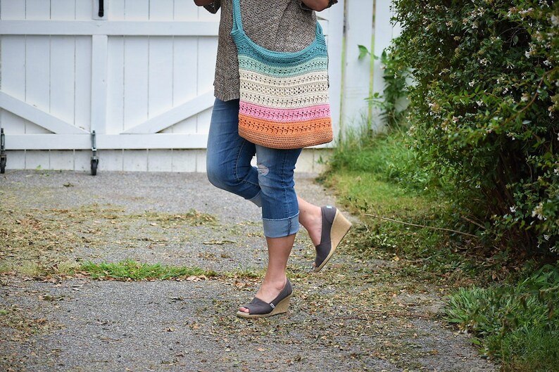 May include: A woman is wearing a pair of blue jeans and a brown sweater. She is carrying a multi-colored crochet tote bag with a teal, white, pink, and orange striped design.