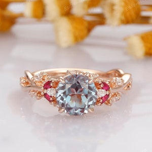 May include: Rose gold ring set featuring a large, round, light blue gemstone as the centerpiece. The ring is accented with smaller red and white gemstones. The band has a delicate, floral design. A second ring with a similar design is also present.