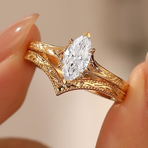 May include: A gold-colored ring set featuring an engagement ring with a marquise-cut diamond and a matching band. The rings have intricate, engraved detailing. The set is held up against a blurred background.